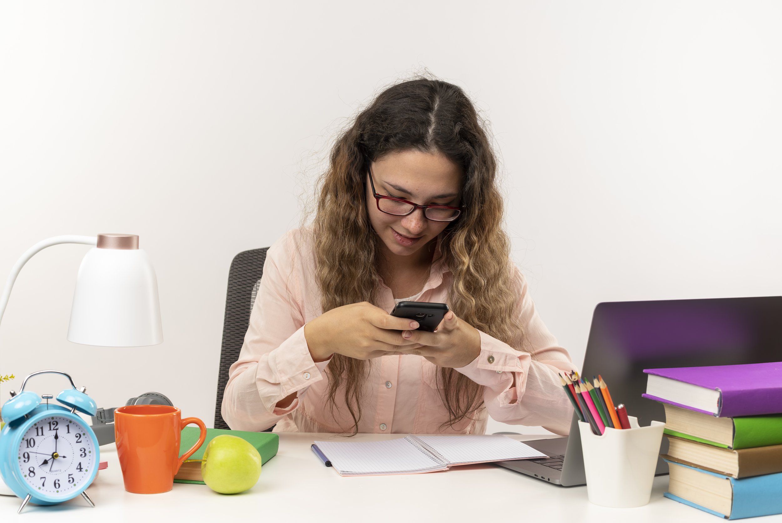 A woman with long curly hair and glasses sitting at a cluttered desk, looking at her phone. The desk has a blue alarm clock, an orange mug, a green apple, a notebook, a laptop, a pen holder with colored pencils, and stacked notebooks.