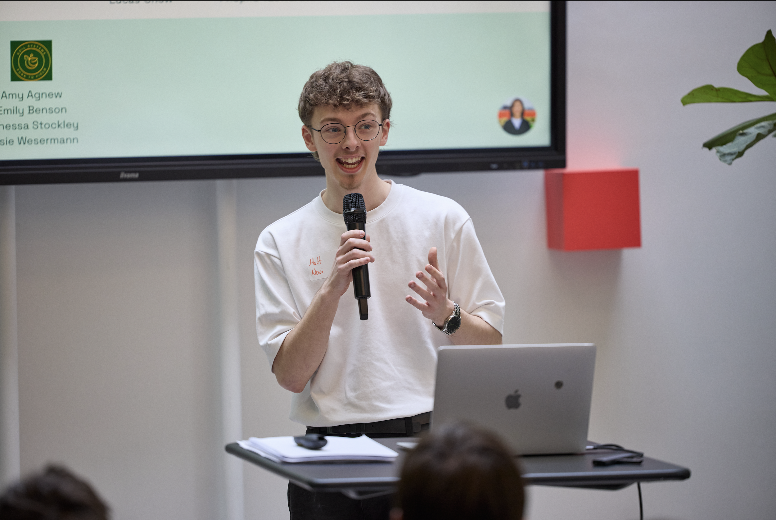 A young man wearing glasses and a white shirt speaking into a microphone in a classroom or presentation setting. A large monitor behind him displays a list of names and a logo, with a laptop and notebook on the table in front of him.