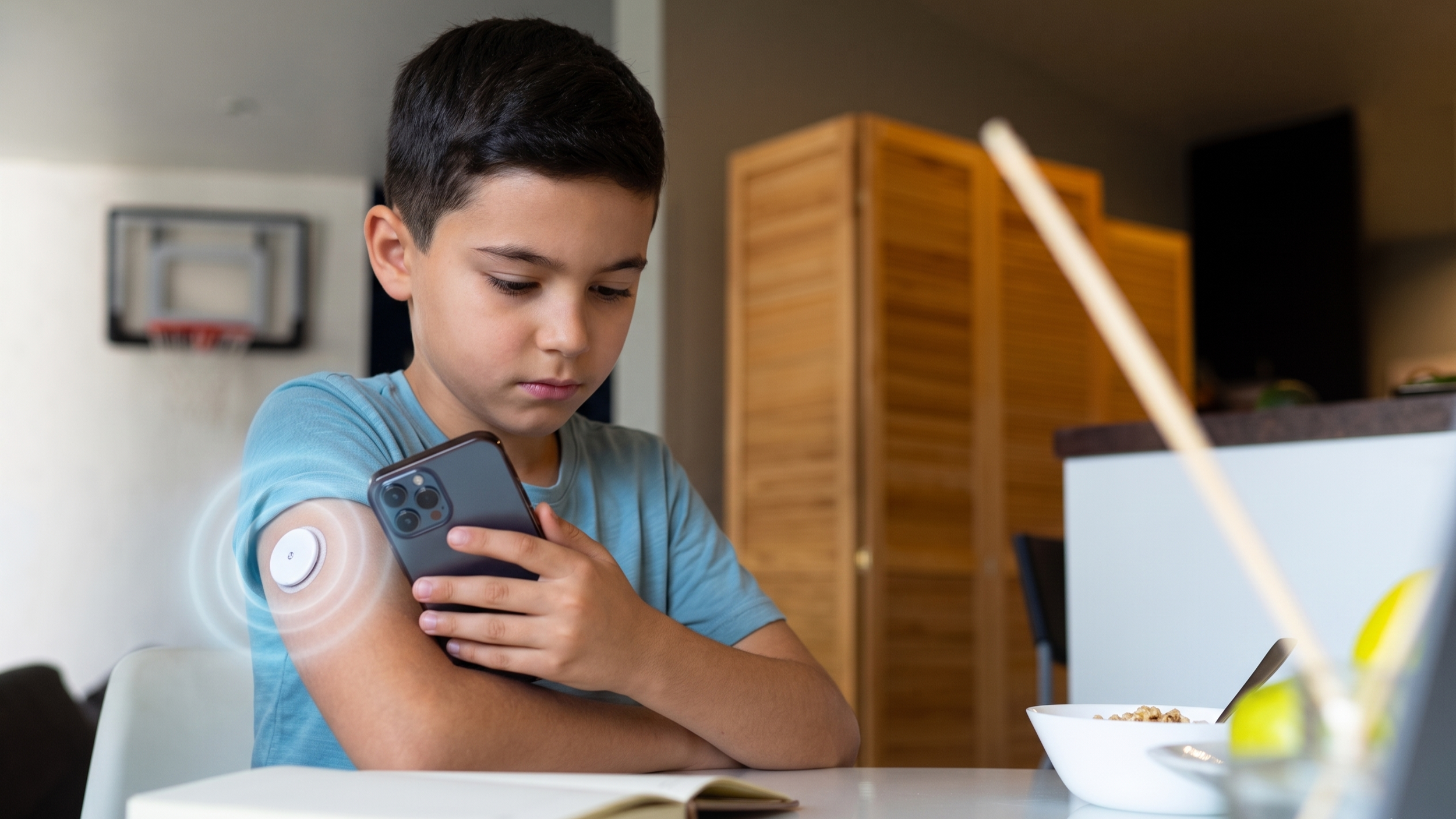 A young boy in a blue shirt checks his blood sugar level using a glucose monitoring device on his arm while holding a smartphone, sitting at a kitchen table with a bowl of cereal nearby.