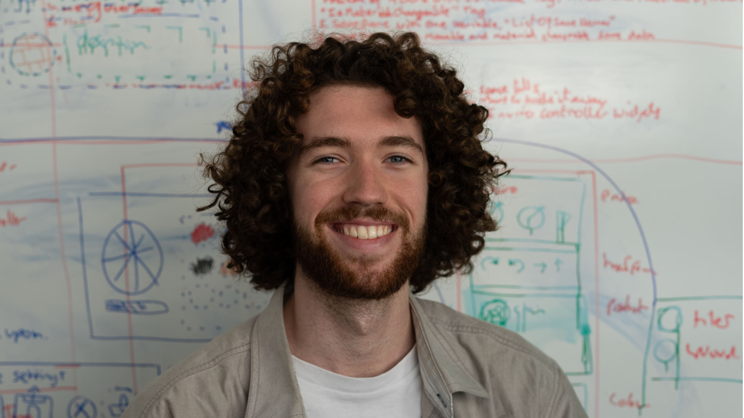 A smiling man with curly brown hair and a beard, standing in front of a whiteboard with colorful diagrams and notes.