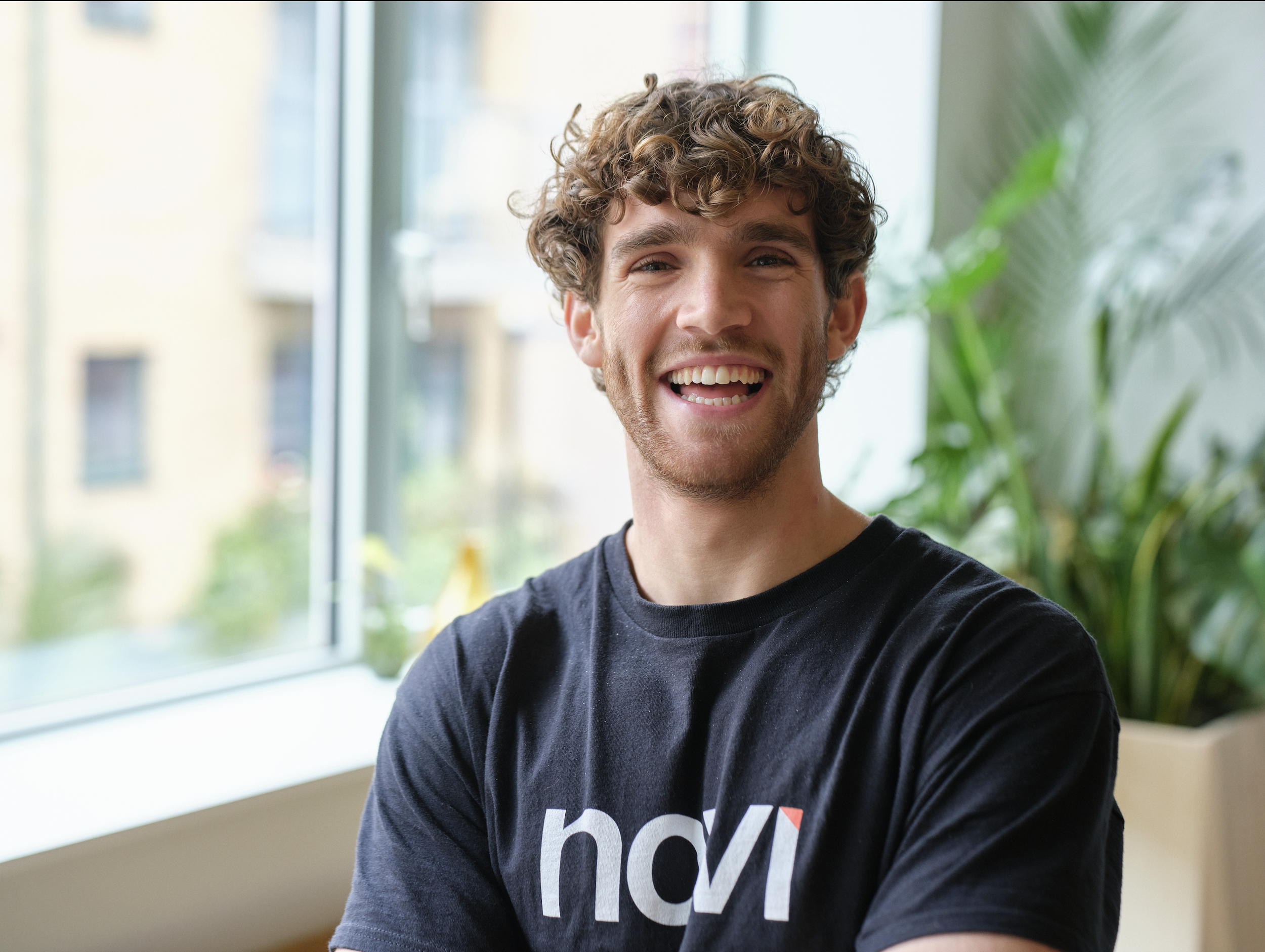 A young man with curly hair smiling and wearing a black t-shirt with white and red lettering, standing indoors near a large window with a blurred background of buildings and greenery.
