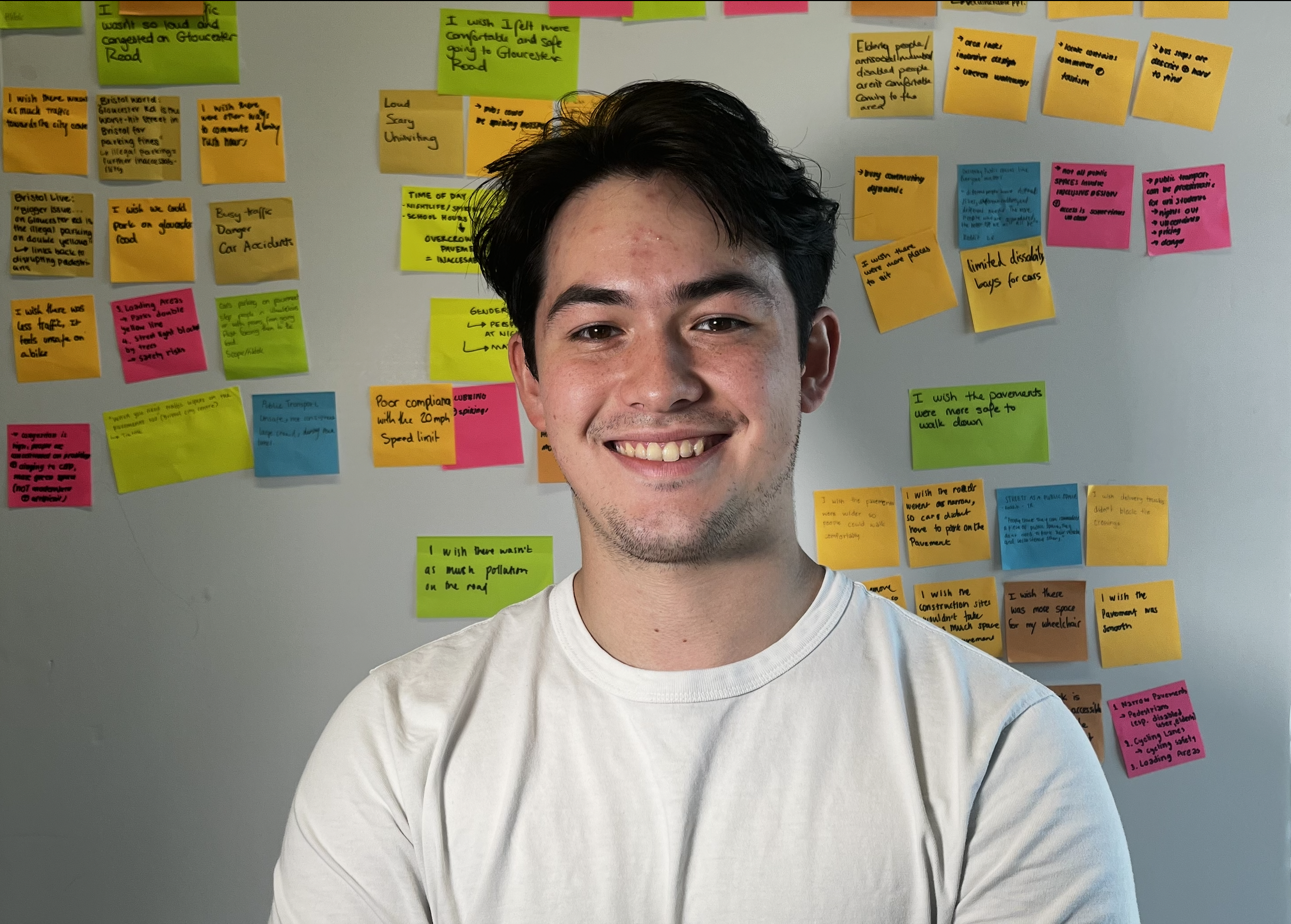 A young man with dark hair and a big smile taking a selfie in front of a wall covered with colorful sticky notes containing handwritten notes and ideas.