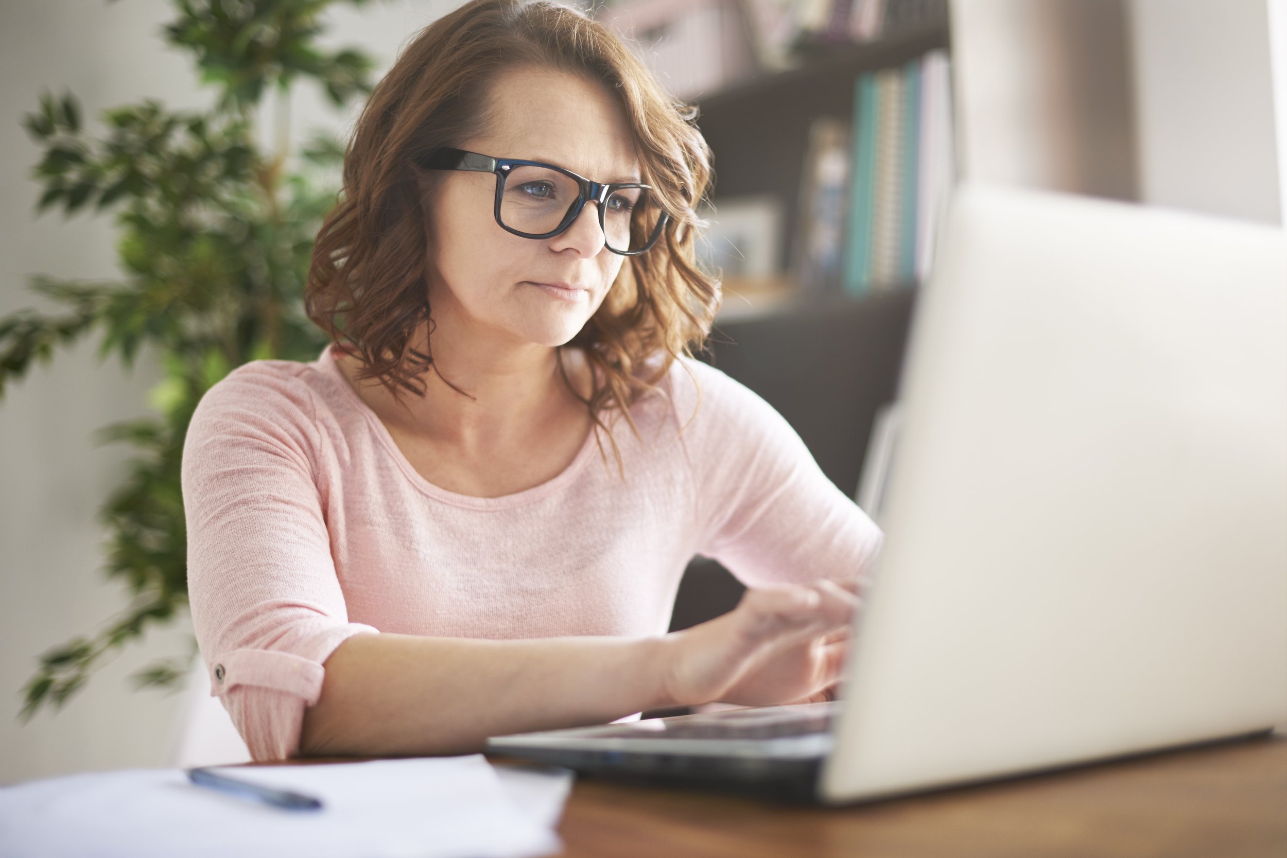 A woman with wavy red hair and black glasses working on a computer in a well-lit room with a plant and bookshelf in the background.